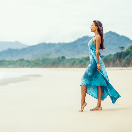 Fashion outdoor photo of gorgeous young sexy lady in long blue dress walking barefoot on the sea shore. Summer sunny day and holiday concept, seascape with girl, beach, beautiful waves, blue water.の写真素材