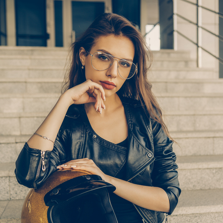 Beautiful young woman in transparent fashion glasses, rock black style leather jacket with glitter gold motorbike helmet look to camera. Hipster urban style girl. Lifestyle outdoor city portrait.の写真素材