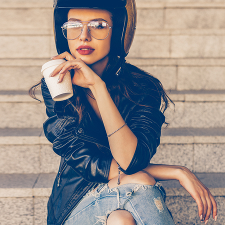 Beautiful young lady in fashion glasses, rock black leather jacket and glitter gold motorbike helmet...