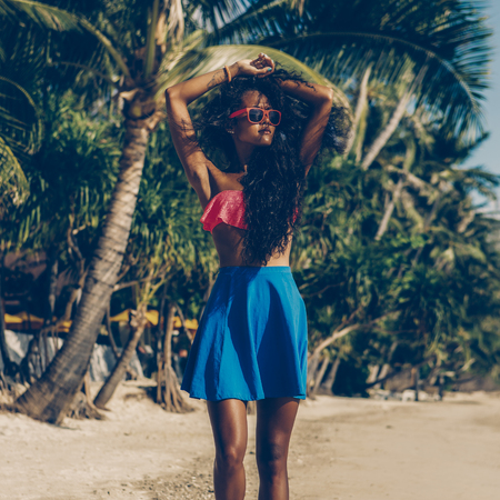 Outdoor lifestyle portrait of black beautiful teenage girl in pink bra and white sneakers. Young hipster woman correct her curly hair. Sunny hot summer day at tropical beach with palms.の写真素材
