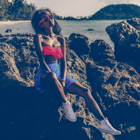 Outdoor lifestyle portrait of a black sexy lady in blue skirt, white converse sneakers, pink bra and sunglasses. Hipster girl sitting at big rock at stone beach. Sunny hot summer day. Swag, fashion.の写真素材
