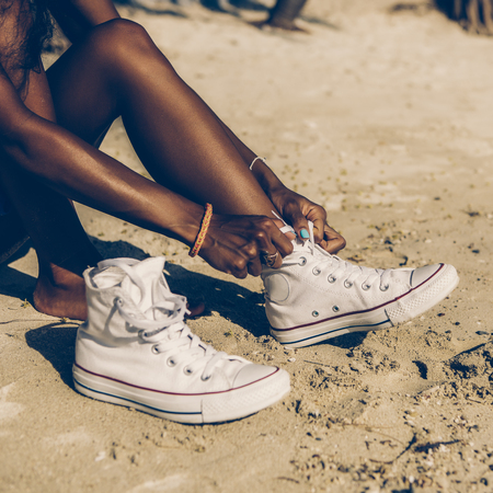 Outdoor lifestyle close-up of the legs of black beautiful teenage girl. Young hipster woman tying laces in her white sneakers. Sunny hot summer day at tropical seashore.の写真素材