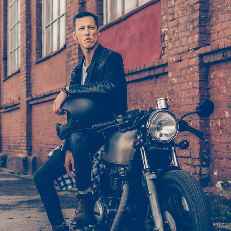 Handsome rider biker guy in black leather jacket sit on classic style cafe racer motorcycle and hold helmet on knee. Bike custom made in vintage garage. Brutal fun urban lifestyle. Outdoor portrait.の写真素材