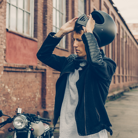 Handsome rider biker man in black leather jacket and denim put on helmet near classic style cafe racer motorcycle. Bike custom made in vintage garage. Brutal fun urban lifestyle. Outdoor portrait.の写真素材