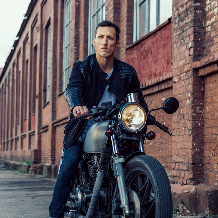 Handsome rider biker guy in leather jacket sit on classic style cafe racer motorcycle and look forward to the future. Bike custom made in vintage garage. Brutal fun urban lifestyle. Outdoor portrait.の写真素材
