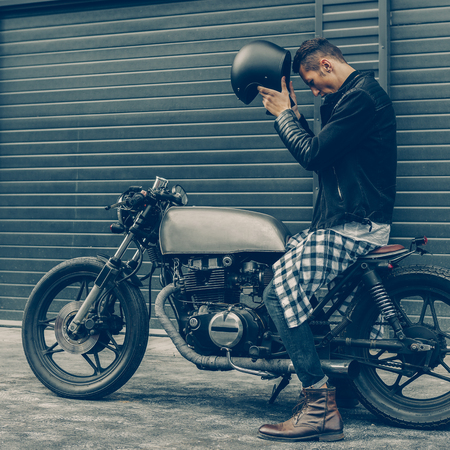 Handsome rider guy take off black biker helmet while sit on classic style cafe racer motorcycle near industrial gates. Bike custom made in vintage garage. Brutal fun urban lifestyle. Outdoor portrait.の写真素材