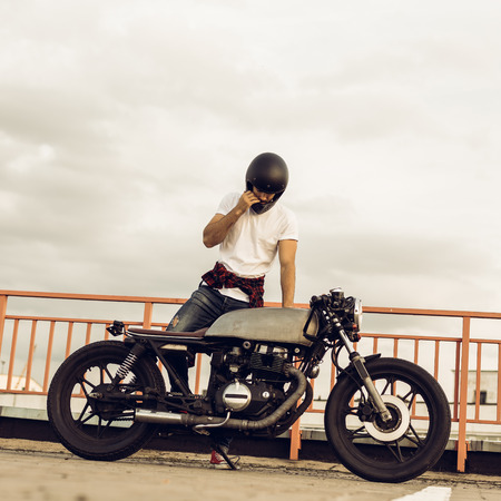 Pretty happy rider man with beard and mustache take off black moto helmet and sit on his classic style biker cafe racer motorcycle. Bike custom made in vintage garage. Brutal fun urban lifestyle.の写真素材