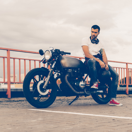 Handsome rider guy with beard and mustache in red sneakers and black moto glasses sit on classic style biker cafe racer motorcycle. Bike custom made in vintage garage. Brutal fun urban lifestyle.の写真素材