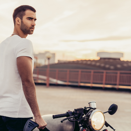Sporty biker handsome rider guy in white t-shirt go travel on classic style cafe racer motorbike on rooftop at sunset. Vintage bike custom made in garage. Brutal urban lifestyle. Outdoor portrait.の写真素材