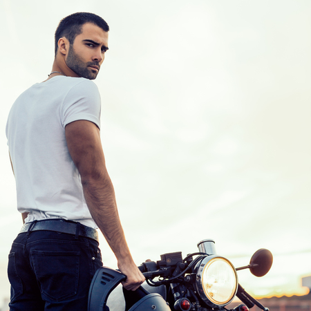 Sporty biker handsome rider man in white t-shirt want to ride his classic style cafe racer motorbike on rooftop at sunset. Vintage bike custom made in garage. Brutal urban lifestyle. Outdoor portrait.の写真素材