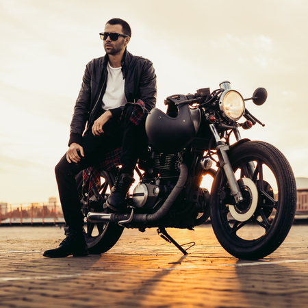 Handsome rider guy with beard and mustache in black leather biker jacket and sunglasses sit on classic style cafe racer motorcycle. Bike custom made in vintage garage. Brutal fun urban lifestyle.の写真素材