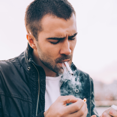 Closeup portrait of a handsome rider man with beard and mustache in black biker jacket, white t-shirt and fashion sunglasses light up cigaret with match on rooftop at sunset. Brutal urban lifestyle.の写真素材