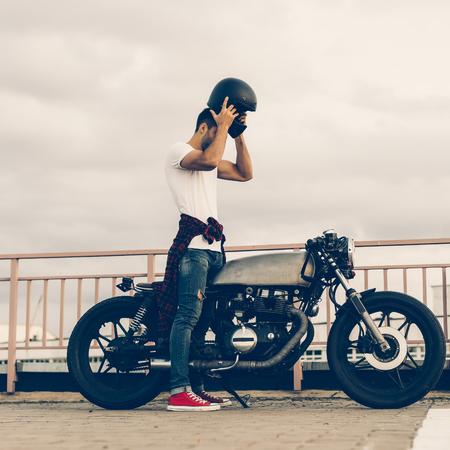 Handsome rider man with beard and mustache posing while put on black moto helmet near classic style biker cafe racer motorcycle. Bike custom made in vintage garage. Brutal fun urban lifestyle.の写真素材