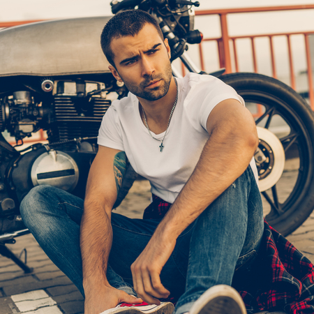 Rider man with beard and mustache in white t-shirt and red sneakers near his classic style biker cafe racer motorcycle. Bike custom made in vintage garage. Brutal fun urban lifestyle.の写真素材