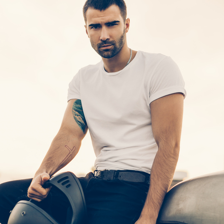 Close up of a handsome rider male in white blank t-shirt look to camera near classic style cafe racer motorcycle at sunset. Bike custom made in vintage garage. Brutal urban lifestyle. Outdoor portrait.の写真素材
