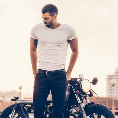 Sporty biker handsome rider man in white blank t-shirt walk away from classic style cafe racer motorbike at sunset. Vintage bike custom made in garage. Brutal urban lifestyle. Outdoor portrait.の写真素材