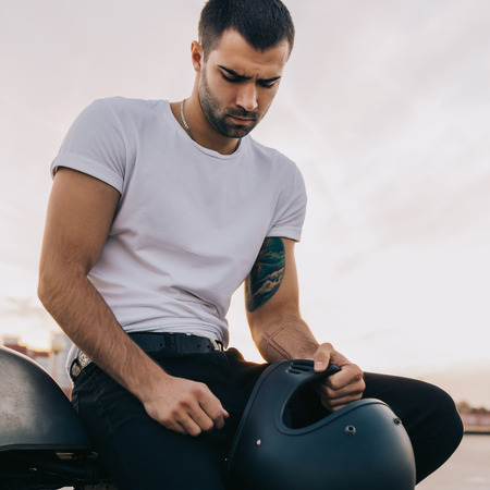 Close up of a handsome rider guy in white blank t-shirt with helmet near classic style cafe racer motorcycle at sunset. Bike custom made in vintage garage. Brutal urban lifestyle. Outdoor portrait.の写真素材