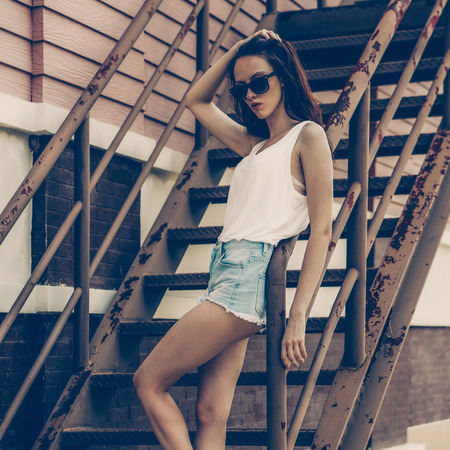 Portrait of young beautiful girl in white t-shirt, short jeans shorts and trendy sunglasses on the stairs. Urban scene, city life. Cute attractive sexy woman touch her long healthy hairs.の写真素材