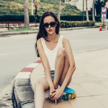 Portrait of slim young beautiful female in white t-shirt, short jeans shorts and trendy sunglasses sit on blue penny skateboard. Urban scene, city life. Cute attractive sexy hipster girl have fun.の写真素材