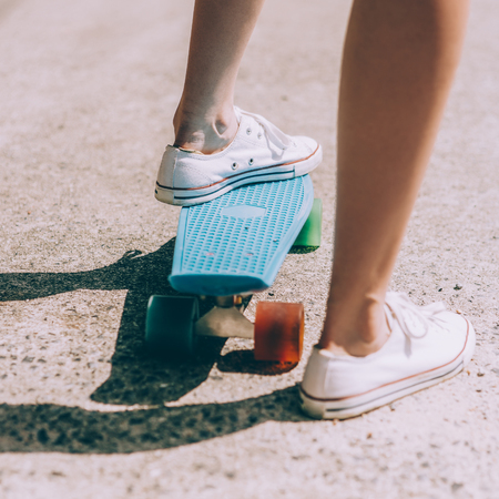 Close up of feet of a beautiful girl in white trendy sneakers rides on blue penny skate board...