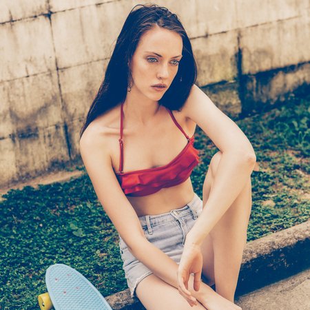 Portrait of pretty beautiful girl in red bra bikini, white sneakers and short denim shorts sit with short skate board. Urban scene, city life. Cute attractive sexy hipster lady looking in front of.の写真素材