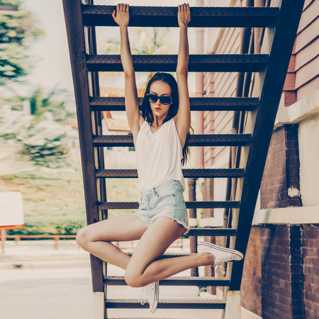 Portrait of sporty beautiful girl in white t-shirt, short jeans shorts and trendy sunglasses pulled on the stairs. Urban scene, city life. Cute attractive sexy hipster lady having fun, fitness, sport.の写真素材