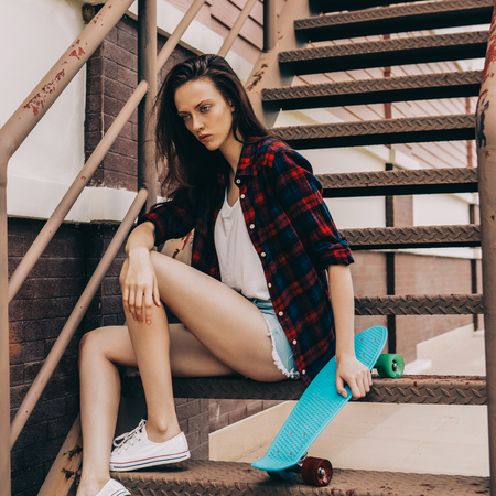 Young beautiful girl in checkered hipster shirt, short jeans shorts and white sneakers sit on the stairs with blue mini skate board longboard skateboard. Urban scene, city life. Cute sexy lady.の写真素材