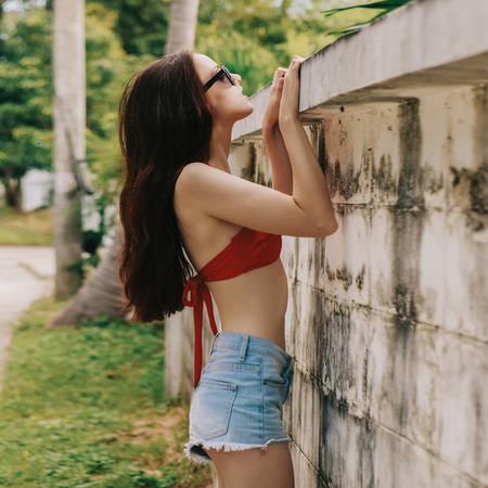 Pretty beautiful woman in red bra bikini, trendy sunglasses and short denim shorts trying climb up a stone wall. Urban scene, city life. Cute attractive sexy hipster lady look over high fence.の写真素材