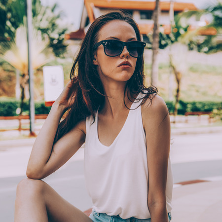 Portrait of sporty beautiful girl in white t-shirt, short jeans shorts and trendy sunglasses sit on blue penny skateboard. Urban scene, city life. Cute attractive sexy hipster lady correct her hairs.の写真素材