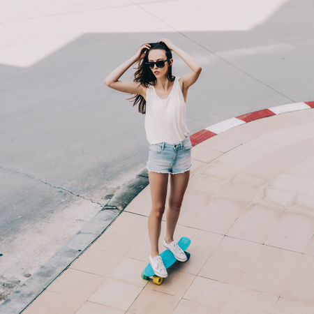 Slim sporty young beautiful girl in white t-shirt, short jeans shorts and trendy sunglasses ride blue penny skateboard and correct hair. Urban scene, city life. Cute attractive sexy hipster lady.の写真素材