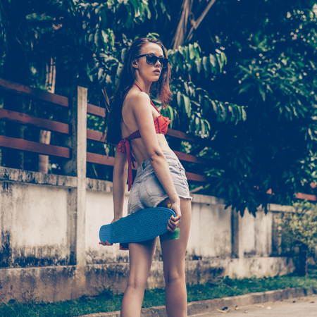 Beautiful girl in red bra bikini, trendy sunglasses, white sneakers and short denim shorts walk with blue penny skate board. Urban scene, city life. Cute attractive sexy hipster lady posing to camera.の写真素材
