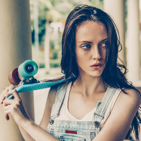 Closeup portrait of sexy beautiful woman in white t-shirt and short denim overall holding blue short skate board. Urban scene, city life. Cute attractive sexy hipster lady standing in front of column.の写真素材