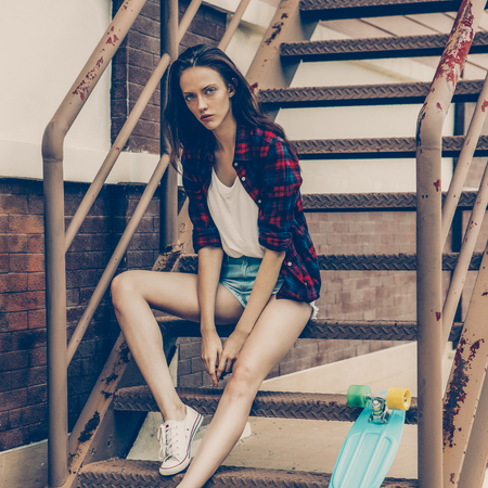 Young beautiful girl in checkered hipster shirt and short jeans shorts and white sneakers sit on the stairs with her blue mini skateboard longboard. Urban scene, city life. Cute lady look to camera.の写真素材
