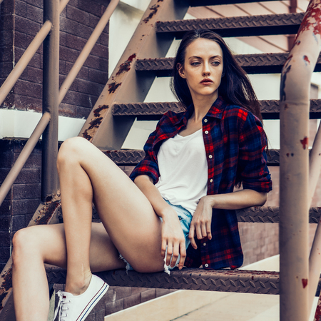 Young beautiful female in checkered hipster shirt and short jeans shorts and white sneakers sit on the stairs with her blue mini skateboard longboard. Urban scene, city life. Woman sexy cute girl.の写真素材