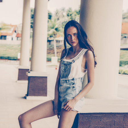 Portrait of slim beautiful girl in white t-shirt and short jeans overall. Urban scene, city life. Cute attractive sexy hipster lady standing in front of a column and posing for the camera.の写真素材