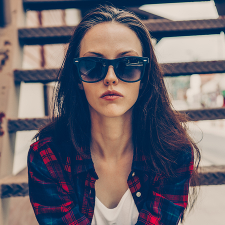 Close-up portrait of young beautiful girl in checkered hipster shirt, short jeans shorts, white sneakers and sunglasses sit on the stairs. Urban scene, city life. Cute sexy lady look to the camera.の写真素材