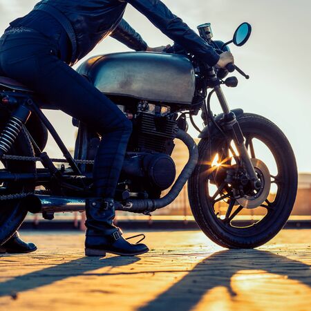 Sexy biker woman in black leather jacket and full face helmet sit on vintage custom caferacer motorbike. Urban roof parking, sunset in big city. Traveling and active hipster lifestyle. Girls power.の写真素材