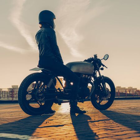 Sexy biker female in black leather jacket and full face helmet sit on vintage custom caferacer motorbike. Urban roof parking, sunset in big city. Traveling and active hipster lifestyle. Girls power.の写真素材