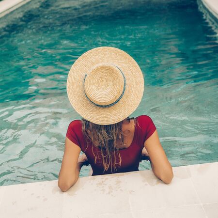 Back of a beautiful young female in sexy red swimsuit and vintage straw hat posing in blue pool water. Hot sunny day. Tropic island vacation hotel. Summer travel girl, active hipster lifestyle.の写真素材