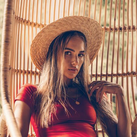 Beautiful young female in fashion sexy red swimsuit and vintage straw hat sit on vogue rattan wooden chair. Hot sunny day. Tropic island vacation. Summer travel girl, active hipster lifestyle.の写真素材