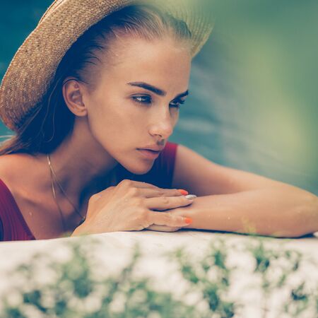 Fashion portrait of a beautiful young female in sexy red swimsuit and vintage straw hat posing in blue pool water. Hot sunny day. Tropic island vacation. Summer travel girl, active hipster lifestyle.の写真素材