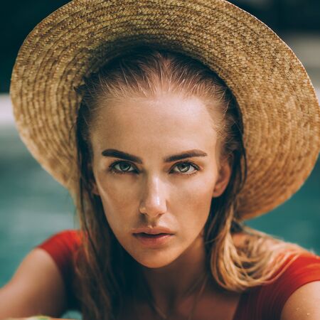 Closeup portrait of a beautiful young woman in sexy red swimsuit and vintage straw hat posing in blue pool water. Hot sunny day. Tropic island vacation. Summer travel girl, active hipster lifestyle.の写真素材