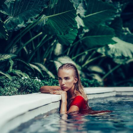 Fashion portrait of a beautiful young female in sexy red swimsuit posing in clear blue pool water. Hot sunny day. Tropic island vacation. Summer travel girl, active hipster lifestyle.の写真素材