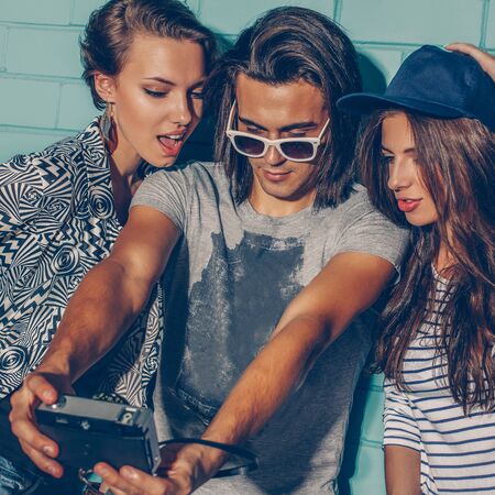 Lifestyle portrait of beautiful best friend hipsters wearing stylish bright outfits and having great time. Handsome boy with two beautiful girls taking selfie self portrait photo with film camera in front of blue brick.の写真素材