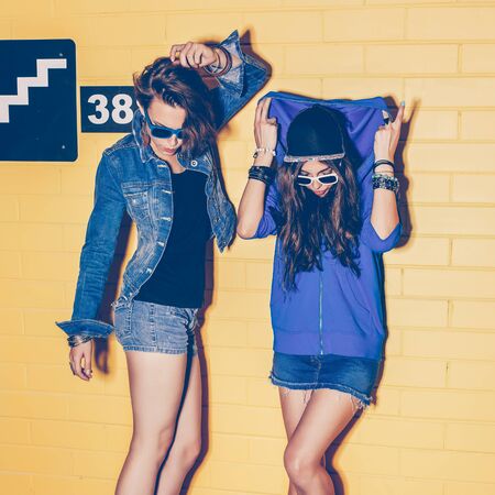 Lifestyle portrait of two beautiful best friends hipster girls wearing stylish bright outfits, denim shorts and sunglasses going crazy and having great time. Standing together near yellow brick wall enjoying day off and have fun.の写真素材