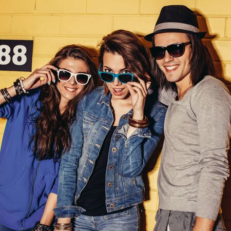 Lifestyle portrait of beautiful adult hipsters wearing stylish bright outfits and sunglasses and having great time. Poses for the camera in front of yellow brick wall enjoying day off and have fun.の写真素材
