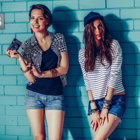 Lifestyle portrait of two beautiful best friends hipster woman wearing stylish bright outfits and denim shorts going crazy and having great time. Standing together near blue brick wall with photo camera and have fun while posing for camera. Young active happy people.の写真素材