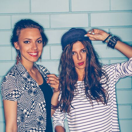 Lifestyle portrait of two beautiful best friends hipster girls wearing stylish bright outfits, denim shorts going crazy and having great time. Standing together near blue brick wall enjoying day off and have fun. Young active happy people.の写真素材
