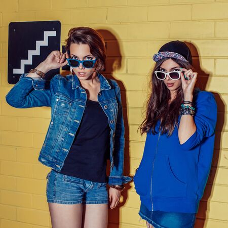 Lifestyle portrait of two beautiful best friends hipster girls wearing stylish bright outfits, denim shorts and sunglasses going crazy and having great time. Standing together near yellow brick wall enjoying day off and correct their glasses.の写真素材