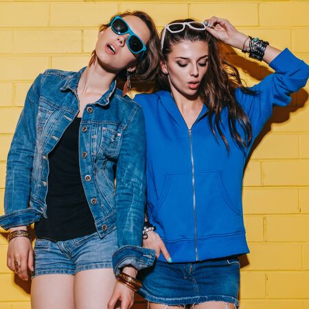 Lifestyle portrait of two beautiful best friends hipster girls wearing stylish bright outfits, denim shorts and sunglasses going crazy and having great time. Standing together near yellow brick wall enjoying day off and posing to the camera.の写真素材